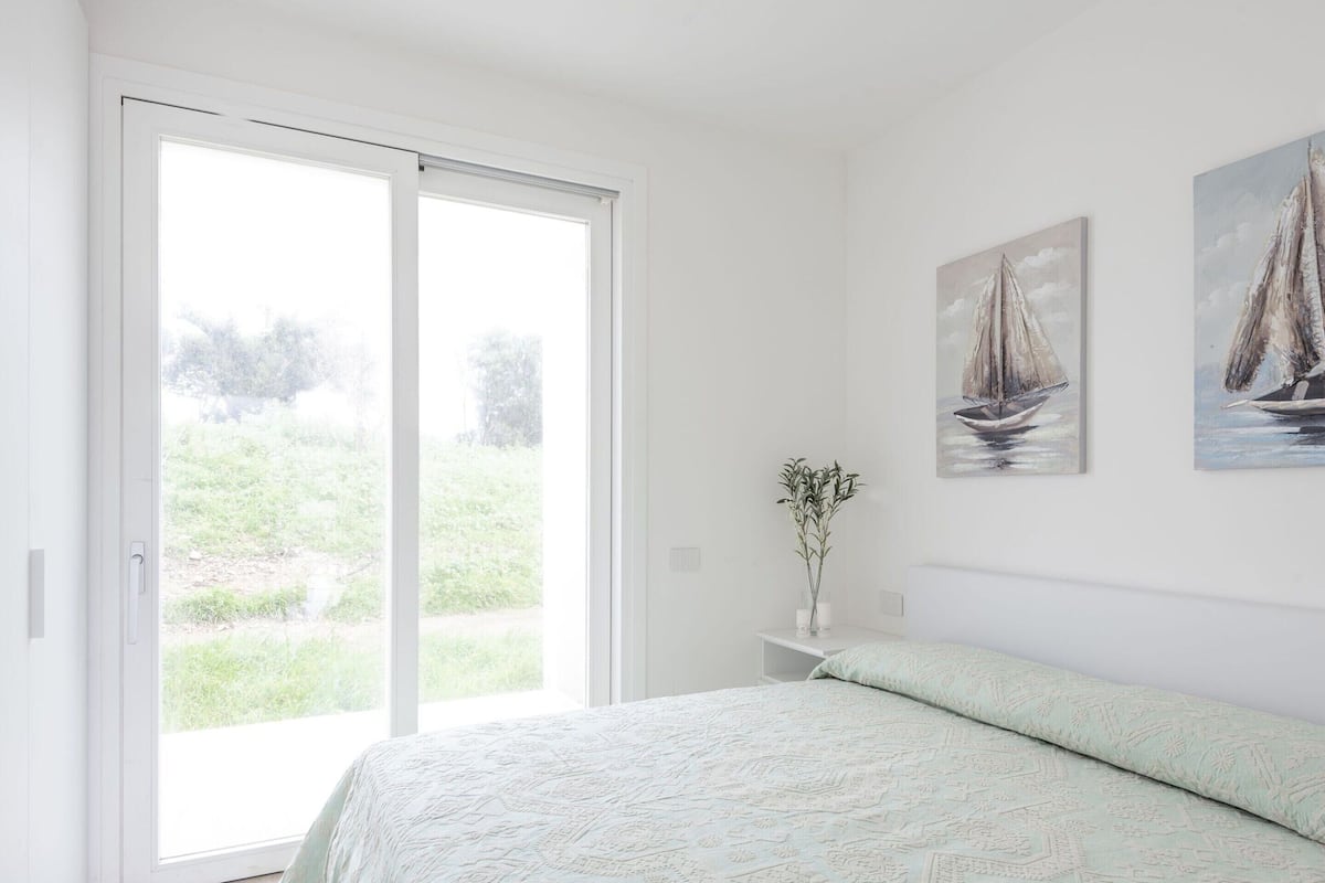 Une chambre avec une grande porte-fenêtre coulissante donnant sur un jardin, un lit recouvert d’une couvre-lit à motifs et deux tableaux représentant des voiliers accrochés au mur.