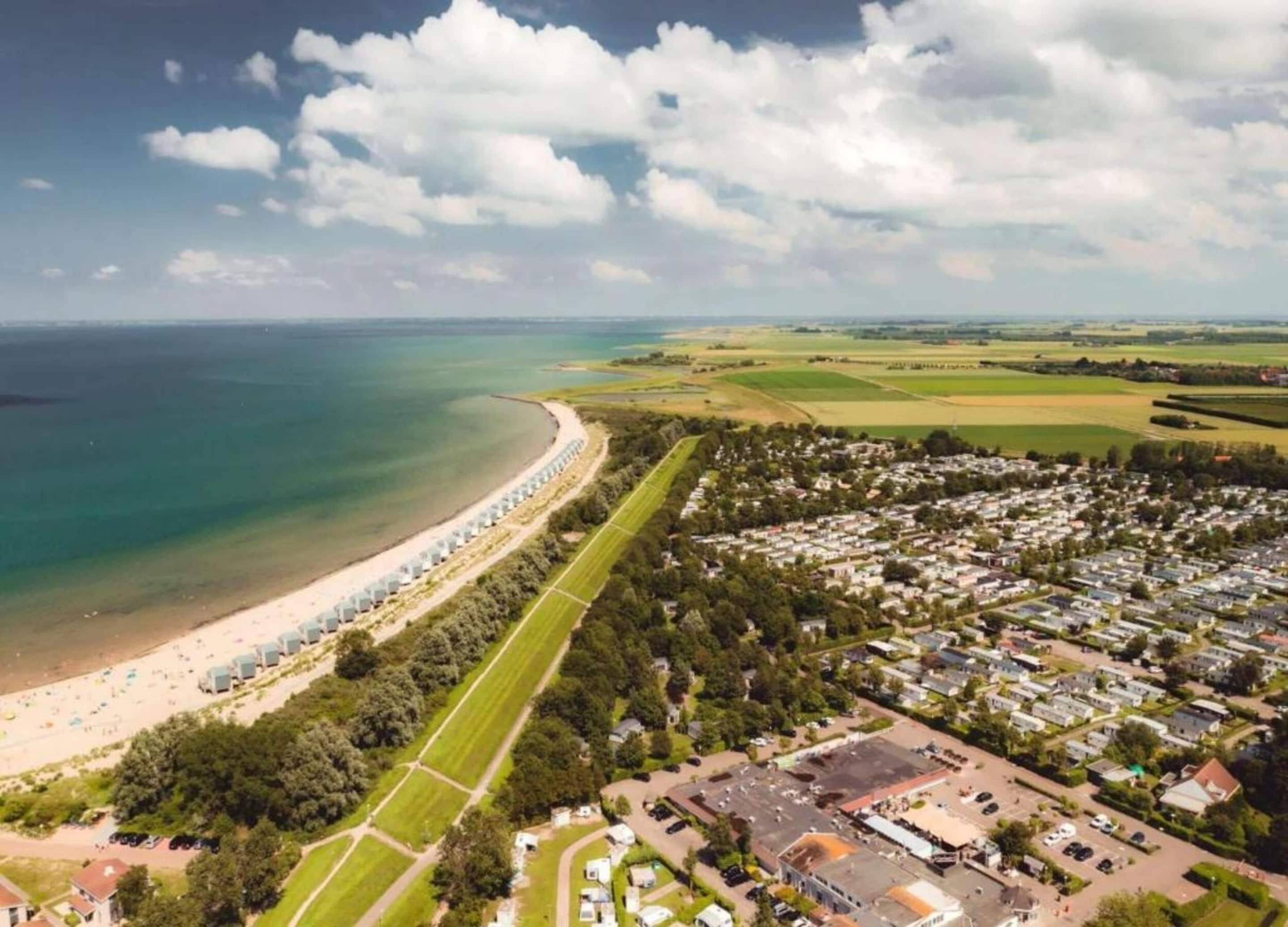 Vue aérienne d’une zone côtière comprenant une plage, une rangée de cabanes de plage et un quartier résidentiel avec des maisons et de la végétation.