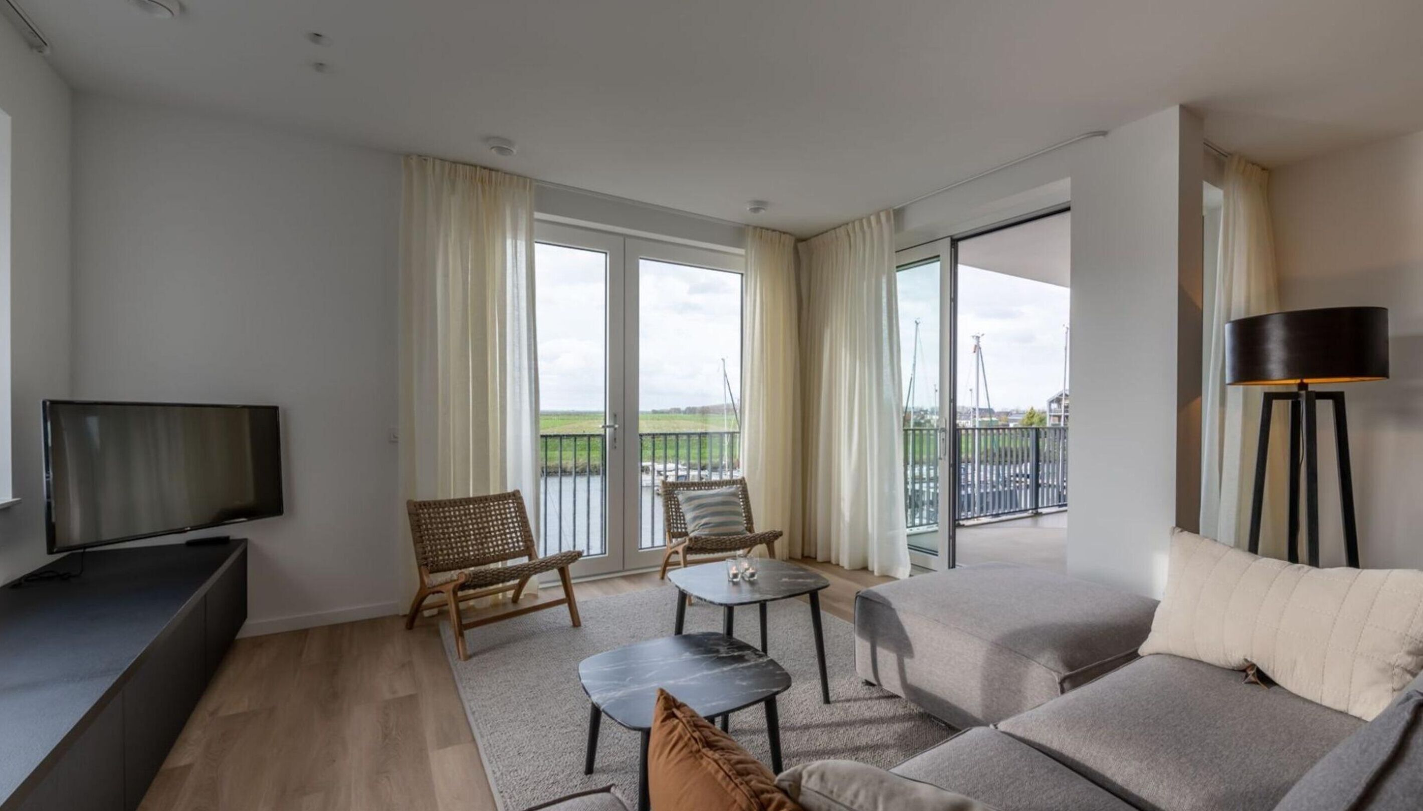 A modern living room with a flat-screen TV, a sofa, a coffee table, and a chair. There is a balcony with a view of the landscape.