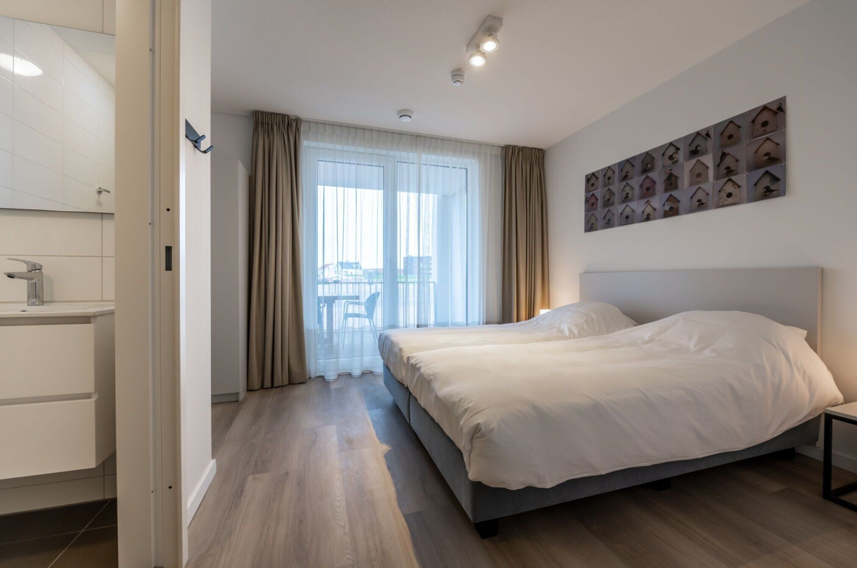 Habitació d'hotel moderna amb un llit gran, un bany amb un lavabo i vistes a la ciutat a través de grans finestres.