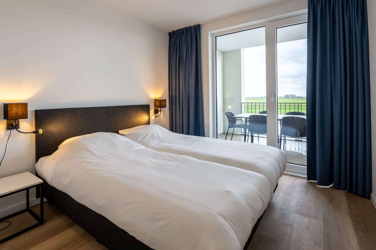 A hotel room with a large bed, two bedside tables, a headboard, and a view of a balcony with outdoor seating.