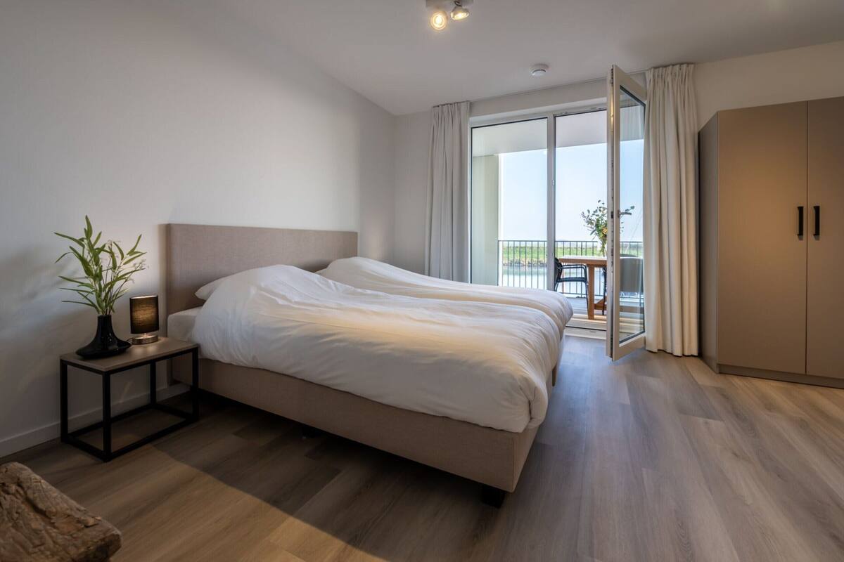 A modern bedroom with a large bed, a bedside table with a lamp, a wardrobe, and a view of the outdoors through a sliding glass door.