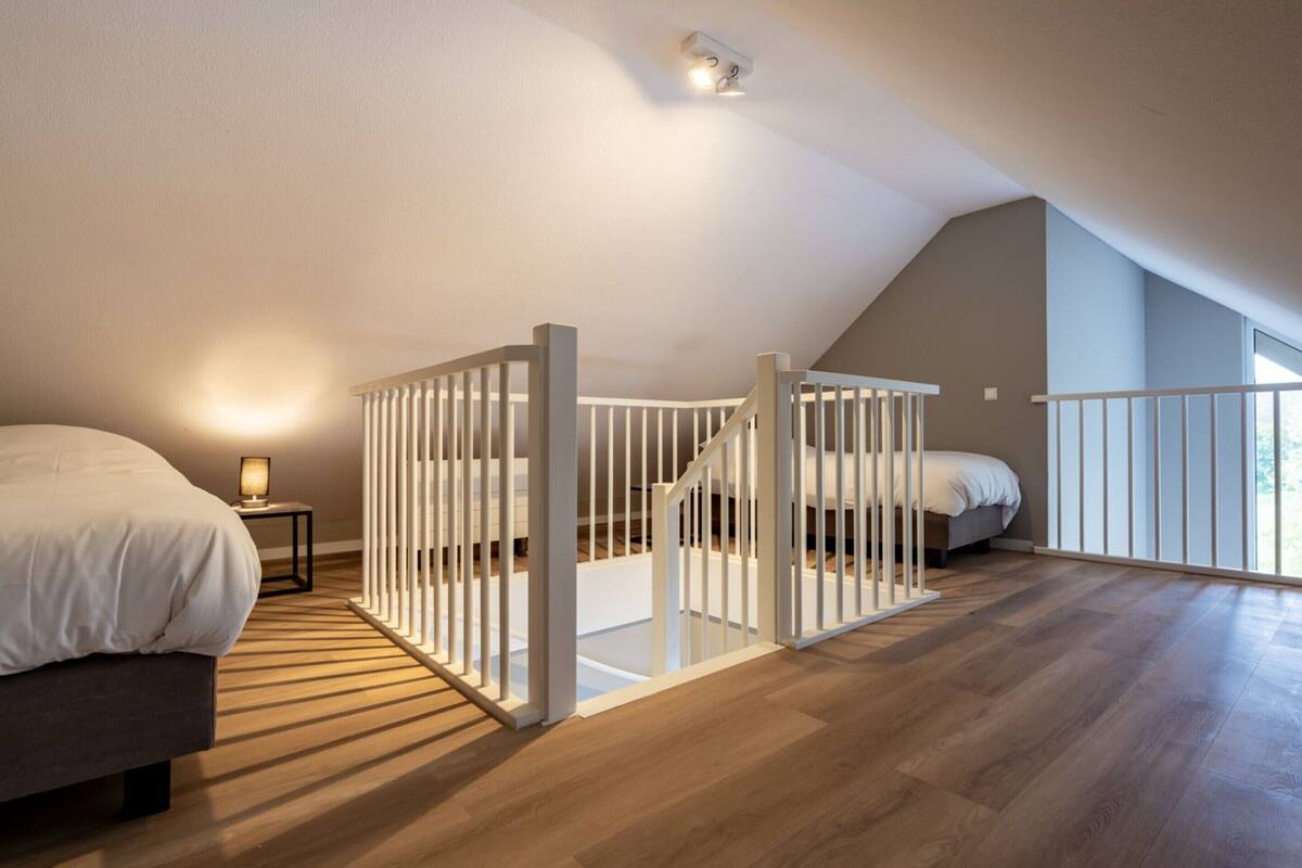 A bedroom with a sloped ceiling, a wooden floor, a bed, a nightstand, and a white railing.