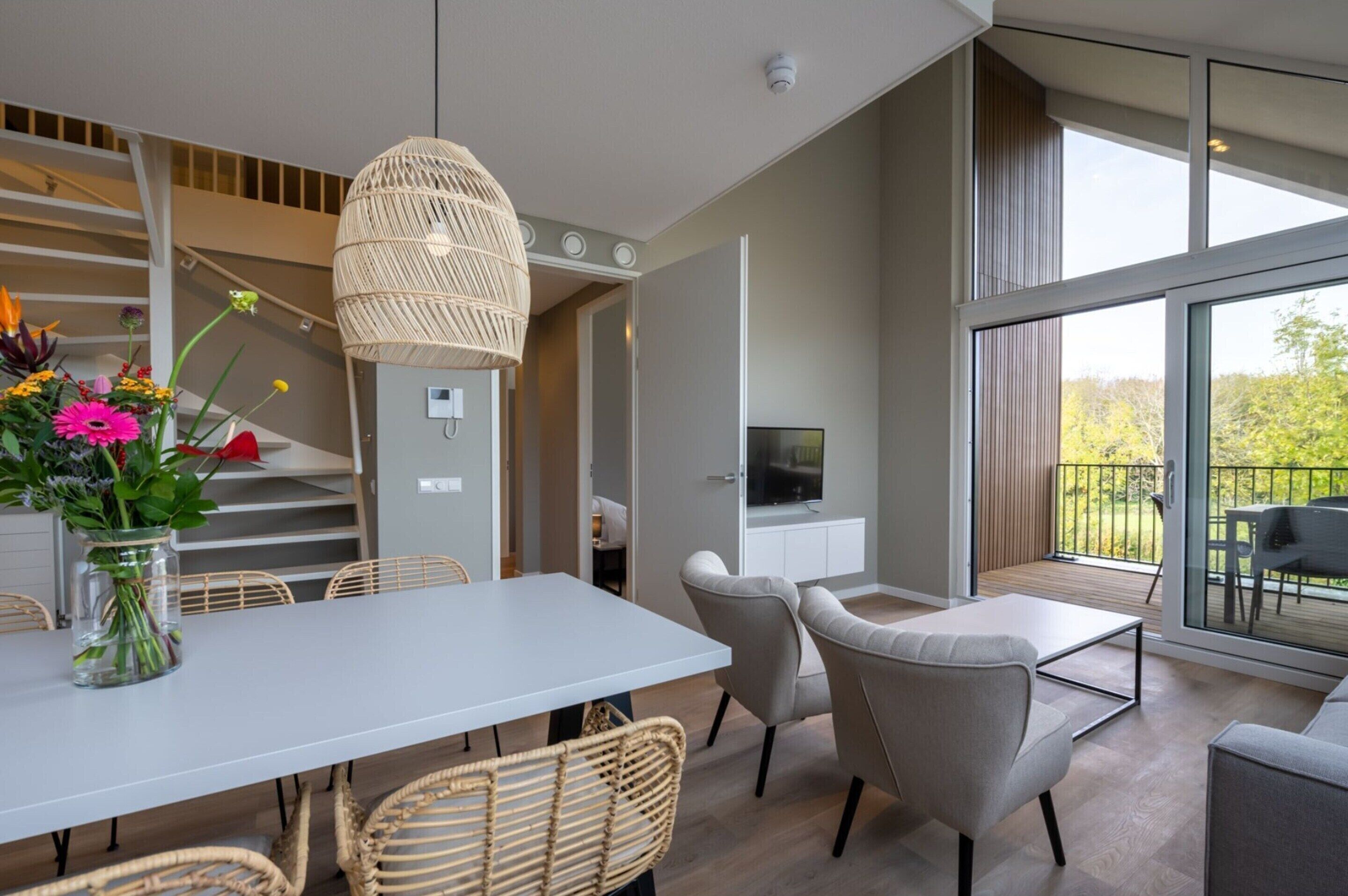 Ein modernes Wohnzimmer mit Essbereich, einem großen Fenster mit Blick auf Grünflächen und einem Fernseher auf einem weißen Schrank.