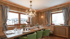 A wooden-paneled dining room with a large table set for four, green chairs, a chandelier, and a view of the outdoors through large windows. - Unique Chalet in the Center of Elmau Near Skilift (Ellmau)