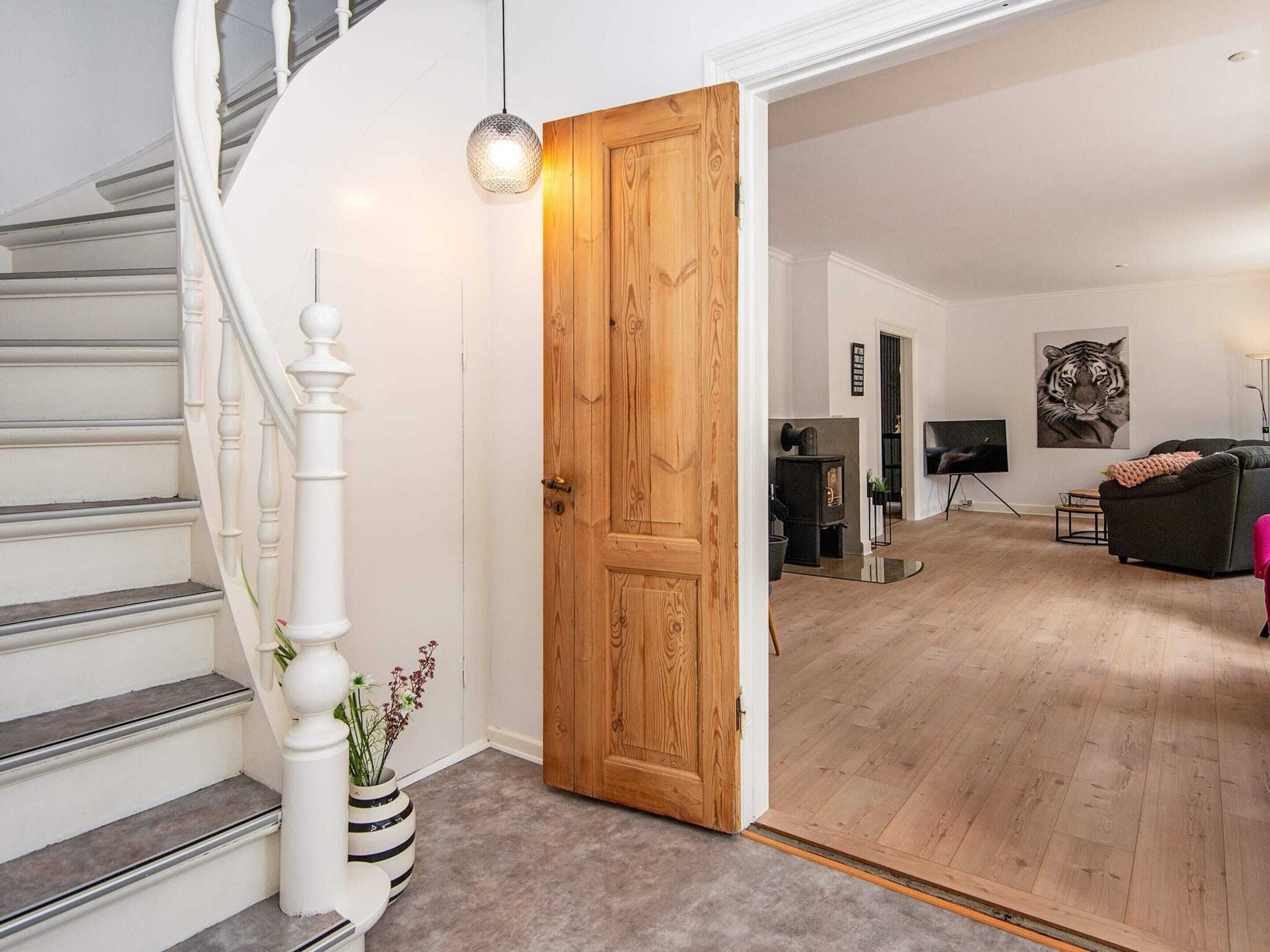 Una escalera con balaustres blancos que conduce a una puerta de madera, una sala de estar con un sofá y una televisión, y una chimenea.
