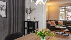 A modern living room with a wooden dining table, a black side table, a sofa with cushions, a floor lamp, and a vase with flowers. - 8 Person Holiday Home in Norre Nebel (Norre Nebel)