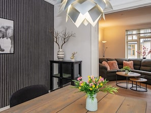 Ein modernes Wohnzimmer mit einem Holzesstisch, einem schwarzen Beistelltisch, einer Couch mit Kissen, einer Stehlampe und einer Vase mit Blumen.