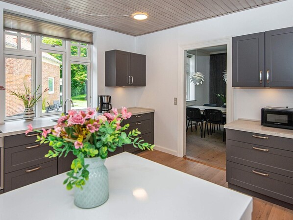 A modern kitchen with dark cabinets, a white countertop, and a vase with flowers. There is a window with a view of a brick building and greenery outside.