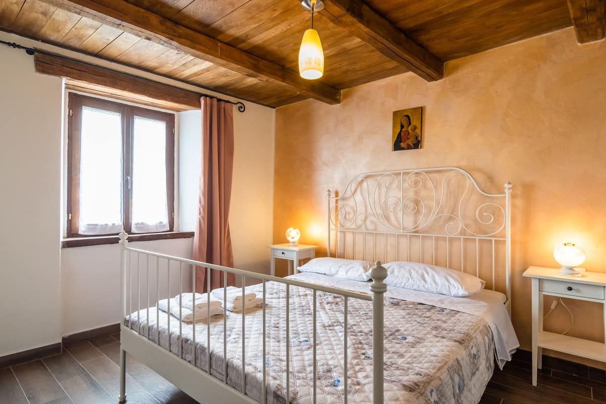 Ein Schlafzimmer mit Holzboden, einem Bett mit Metallkopfteil, zwei Nachttischen mit Lampen, einem Fenster mit Vorhängen und einem Bild an der Wand.