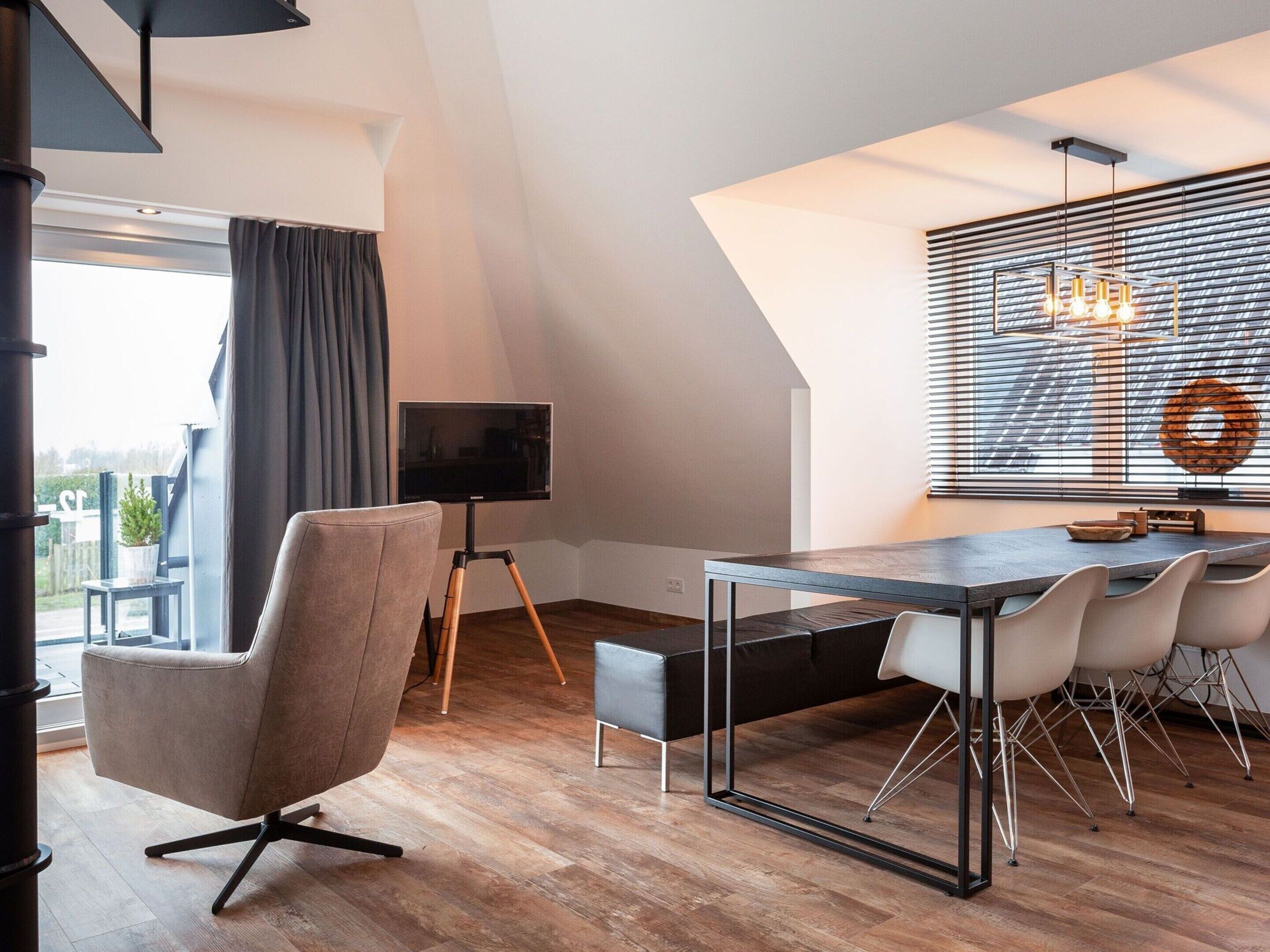 A modern living room with a wooden floor, a dining table with chairs, a TV on a tripod, and a large window with blinds.
