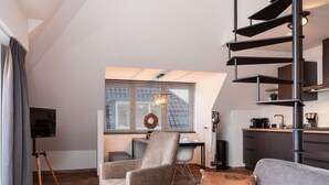 A modern living room with a grey sofa, a coffee table, and a TV on a tripod stand. There is a staircase with a black railing and a kitchen area in the background. - Apartment Located Directly on the Sneekermeer (Offingawier)
