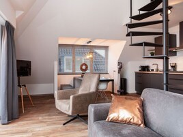 Uma sala de estar moderna com um sofá cinzento, uma mesa de centro e uma TV apoiada em um suporte de tripé. Uma escada com corrimão preto e uma área de cozinha podem ser vistas ao fundo.