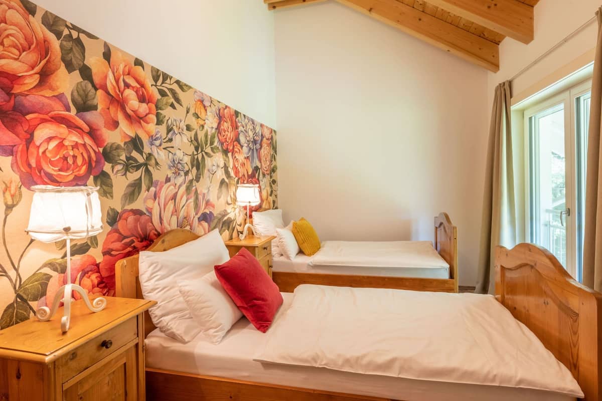 A room with two wooden beds, a floral headboard, a nightstand with a lamp, and a window with curtains.