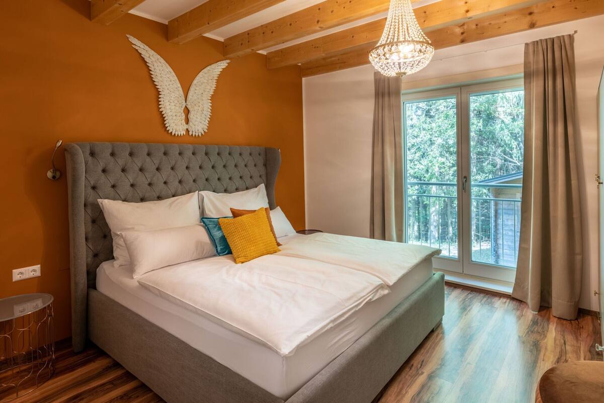 A bedroom with a large bed, wooden ceiling beams, a chandelier, and a window with curtains.