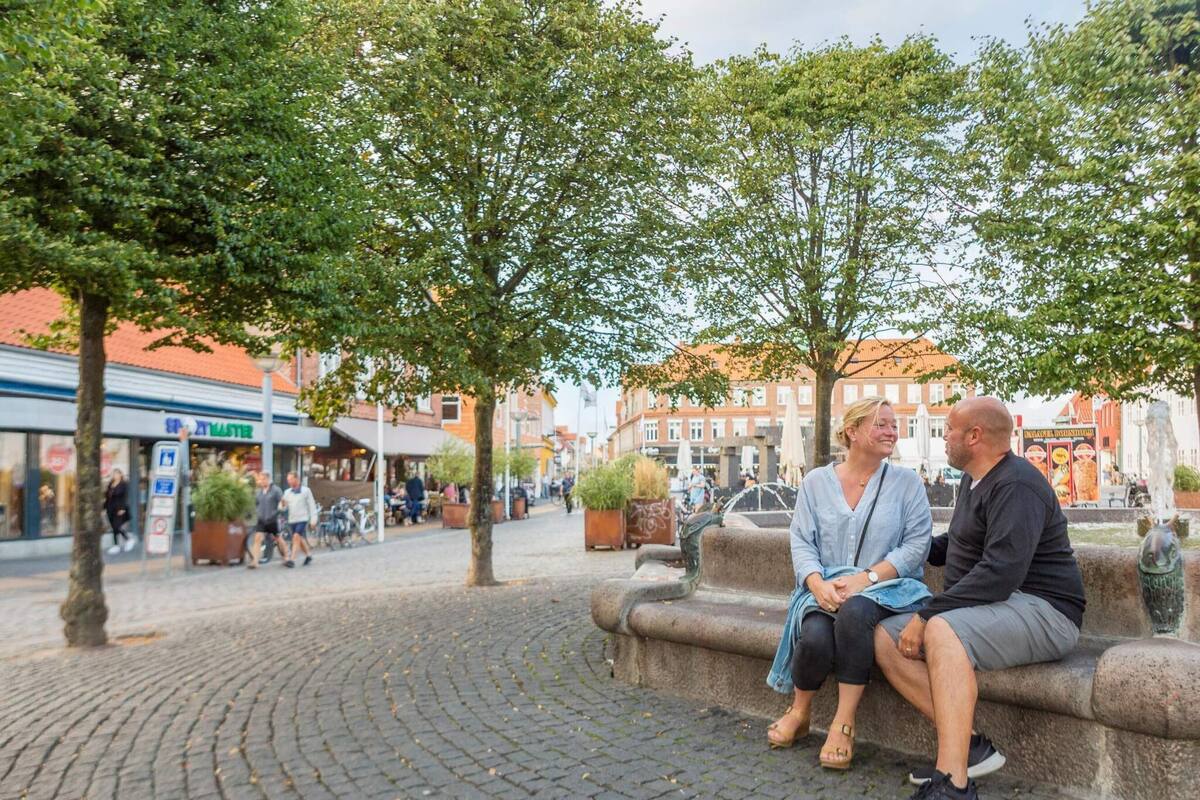 A couple sitting on a stone bench in a cobblestone square with trees and buildings in the background.