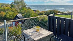 Une terrasse en bois avec une table et des chaises, donnant sur un paysage côtier avec vue sur la mer et un petit bâtiment en arrière-plan.