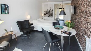 Une chambre d’hôtel moderne comprenant un lit, une table à manger avec des chaises, un fauteuil en cuir noir, une petite table d’appoint avec une lampe et un mur en pierre.