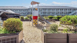 Un terrain de jeux avec un toboggan et une structure d’escalade, entouré de gravier et de clôtures en bois, avec un bâtiment visible au fond.