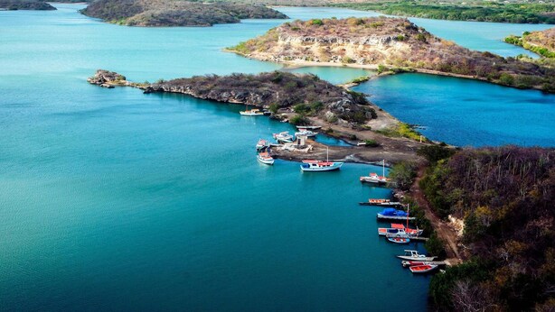Una zona costera con barcos amarrados en un puerto deportivo, rodeada de colinas y un cielo despejado.