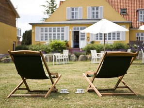 Two wooden lounge chairs with green cushions in a grassy area in front of a yellow building with white shutters.