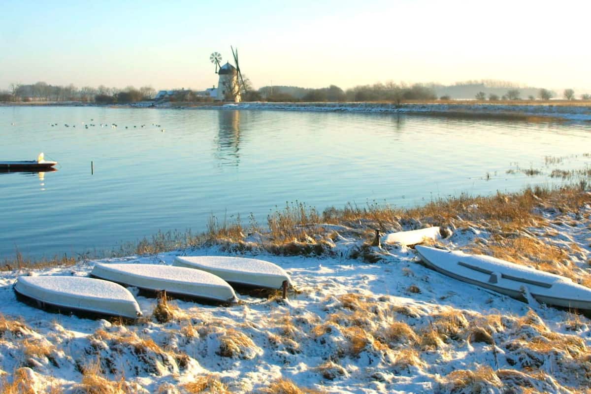 A serene lakeside scene with a windmill in the distance, snow-covered ground, and boats on the shore.
