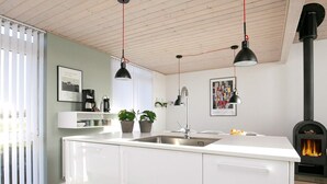 Een moderne keuken met witte kastjes, een centraal kookeiland met een spoelbak, hanglampen, een zwarte kookplaat en een raam met zonwering.