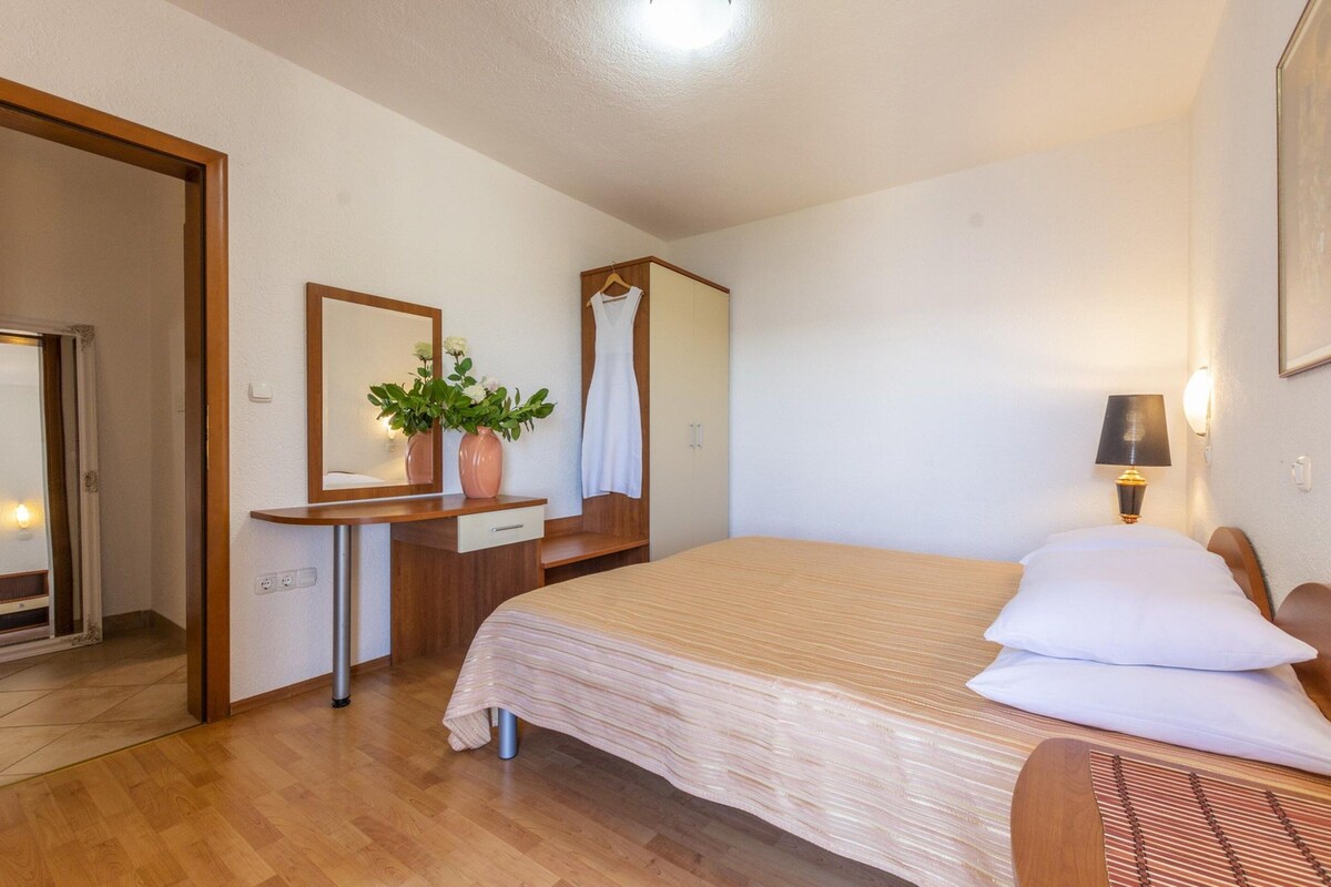 A hotel room with a bed, a desk, a mirror, a plant, and a hanging dress.