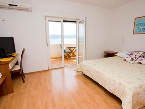 A bedroom with a bed, a desk with a computer, a chair, and a sliding glass door leading to a balcony with a table and chairs.