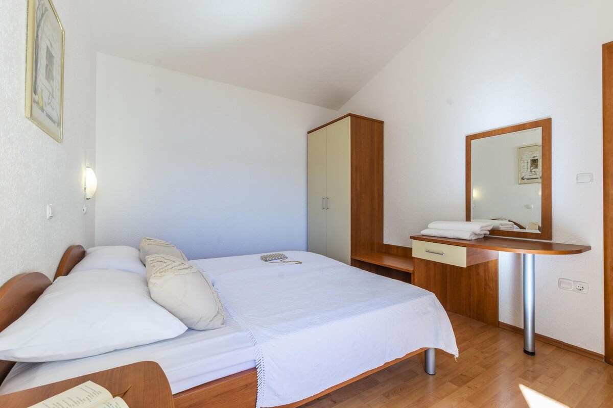 A bedroom with a bed, a wooden wardrobe, a bedside table with a mirror, and a wall-mounted light.