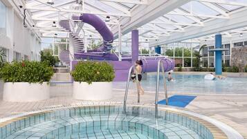 An indoor swimming pool with a slide, surrounded by potted plants and a covered area with a glass roof.