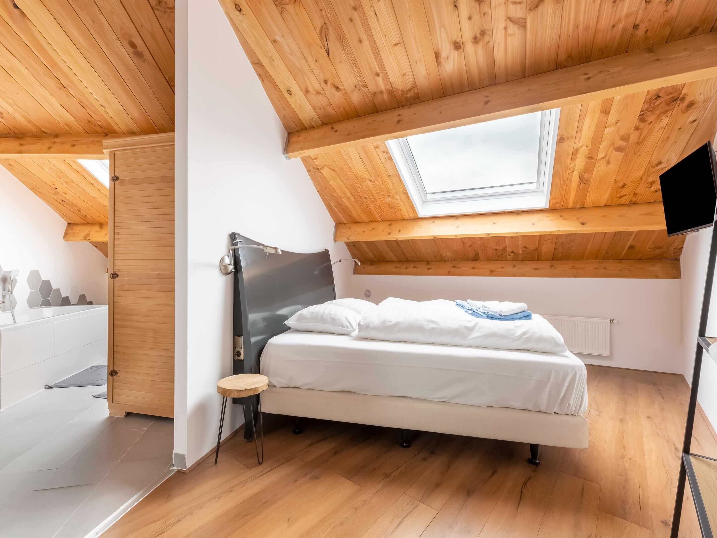 Un dormitorio con tragaluces, vigas de techo de madera, una cama con ropa de cama blanca y un taburete pequeño.