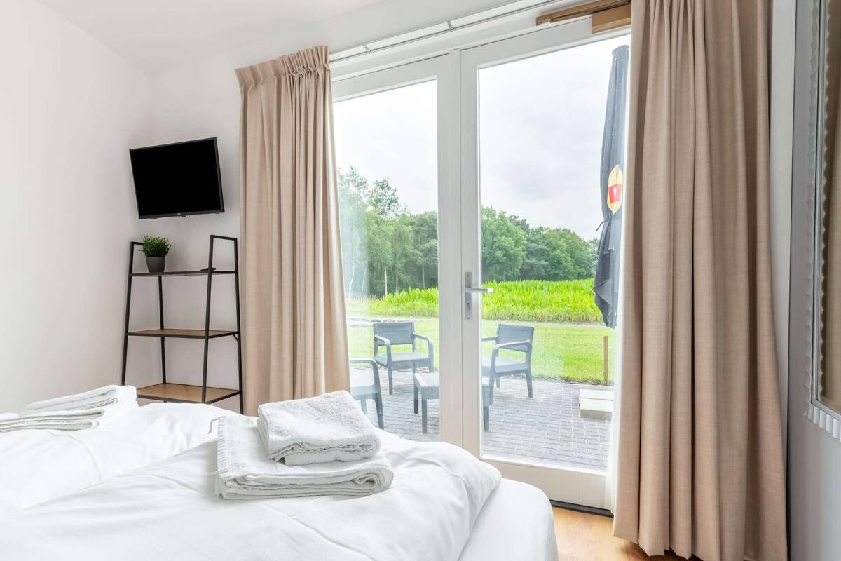 A hotel room with a bed, a TV mounted on the wall, a small shelf with a plant, and a view of a garden through a sliding glass door.