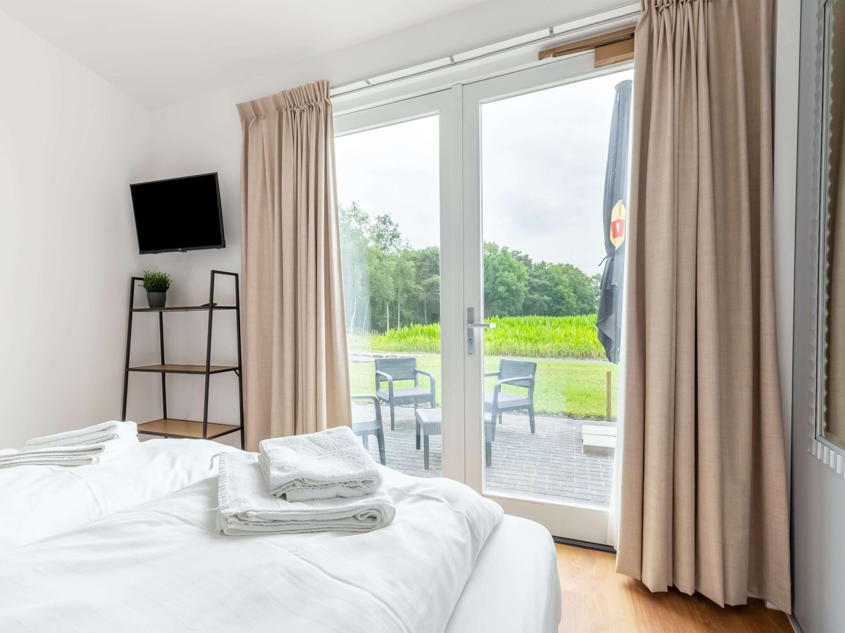 A hotel room with a bed, a TV mounted on the wall, a small shelf with a plant, and a view of a garden through a sliding glass door.