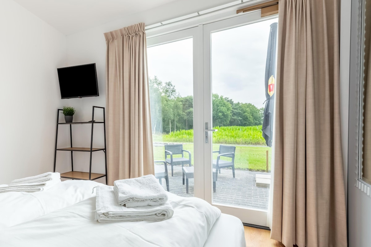 A hotel room with a bed, a TV, a plant, and a view of the garden through the door.