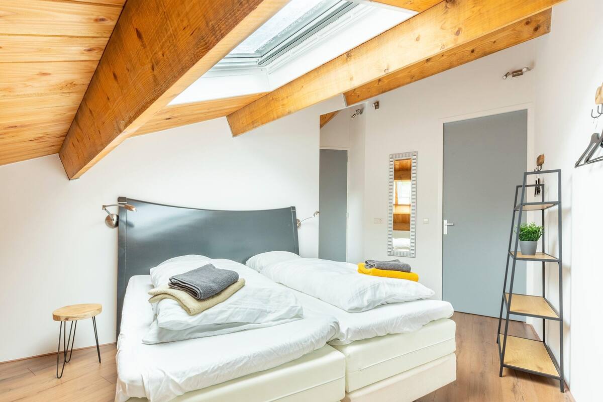A modern bedroom with a skylight, a bed with neatly folded towels, a small stool, and a wooden ladder shelf.