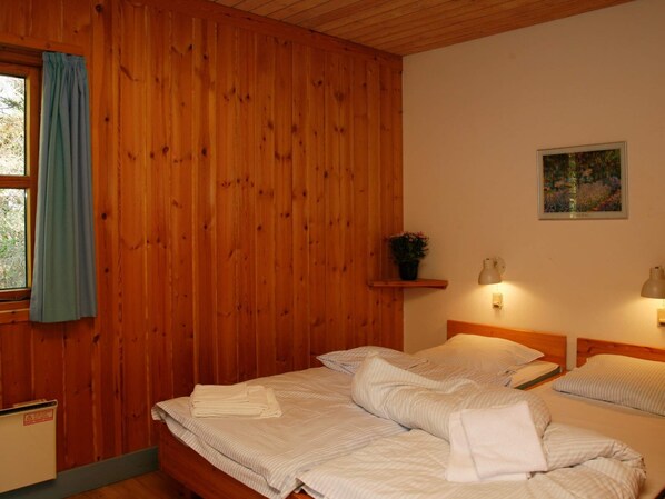 A room with wooden walls, two beds with striped bedding, a window with blue curtains, a small shelf with a plant, and wall-mounted lamps. - 6 Person Holiday Home on a Holiday Park in Vaeggerlose (Vaeggerlose)