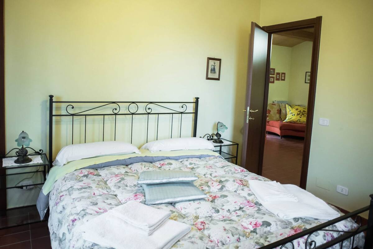 A bed with a floral bedspread, two pillows, and a folded white towel. There is a nightstand with a lamp and a framed picture on the wall.