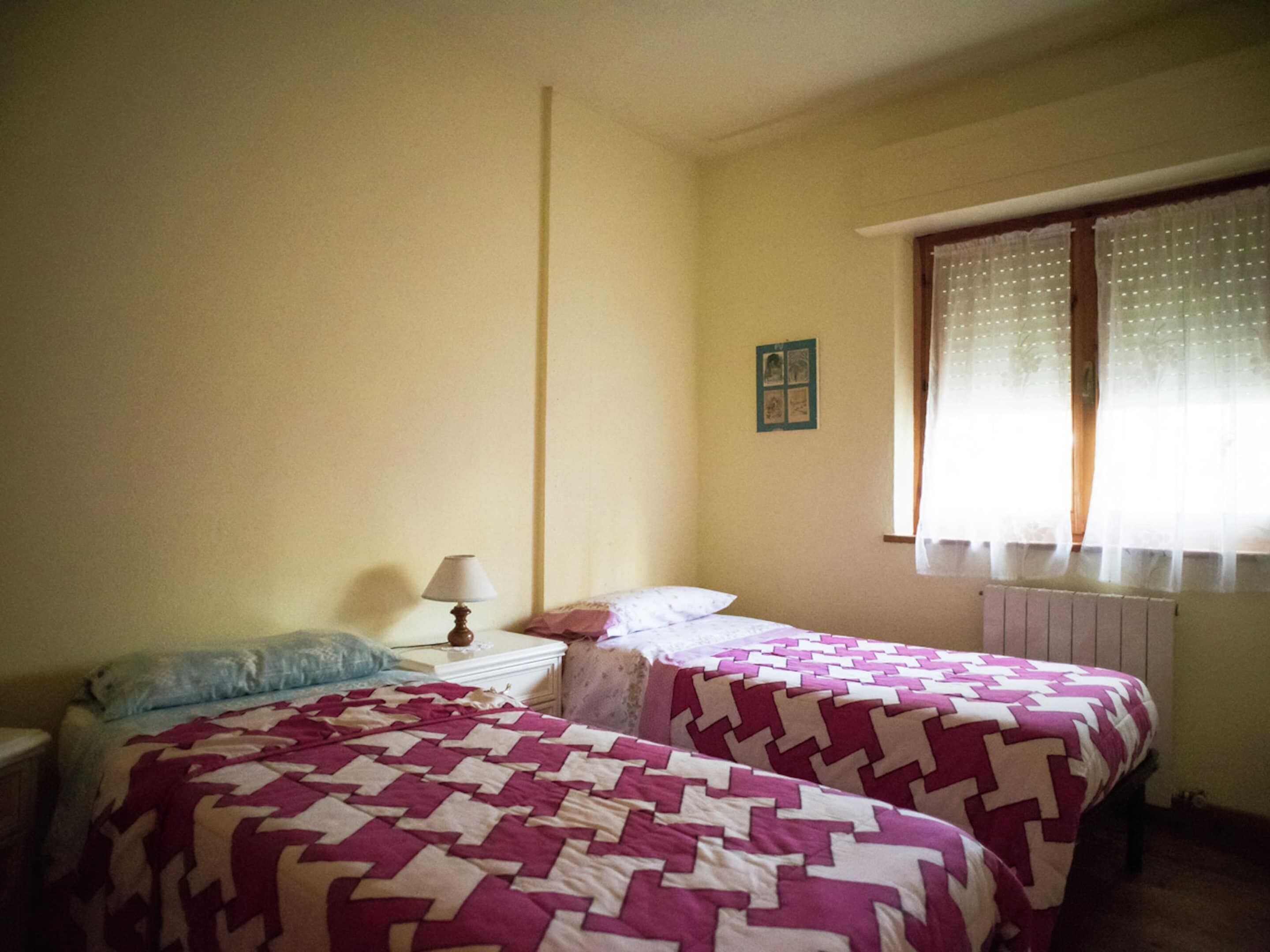 A room with two beds, a nightstand, a lamp, a window with curtains, and a wall-mounted picture.