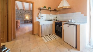 A compact kitchen with a stove, oven, and sink. There's a wooden door leading to a dining area with a stone wall and a lamp. - Chic Farmhouse in Montalto di Castro With Terrace (Montalto di Castro)