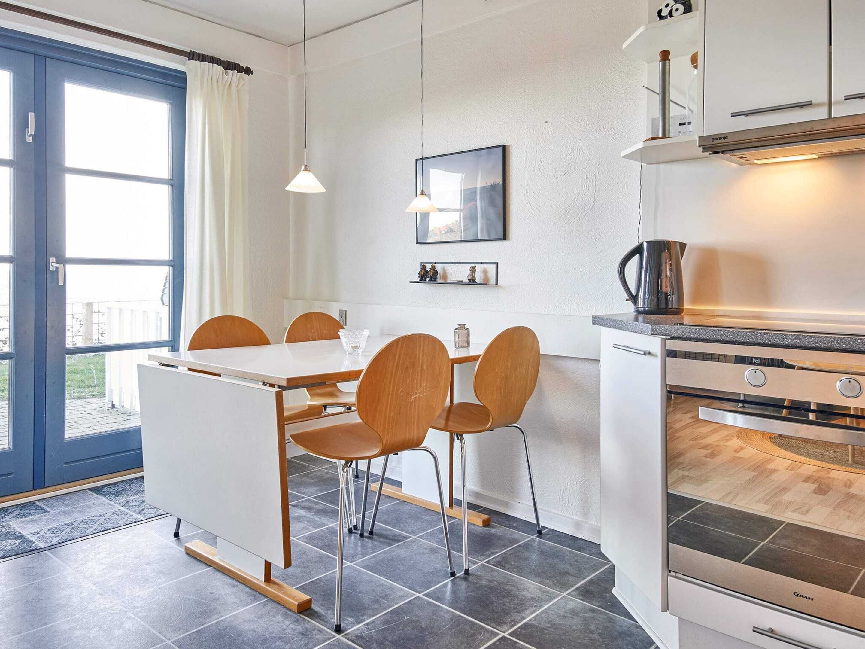 A modern kitchen with a built-in oven, a dining area with a white table and wooden chairs, and a large window with curtains.