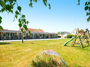 Un edificio con techo de tejas rojas, un patio cubierto con mesas y sillas, y un área de juegos con tobogán y columpios.