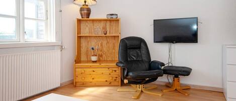 A living room with a wooden cabinet, a black leather chair, a TV on the wall, and a radiator.