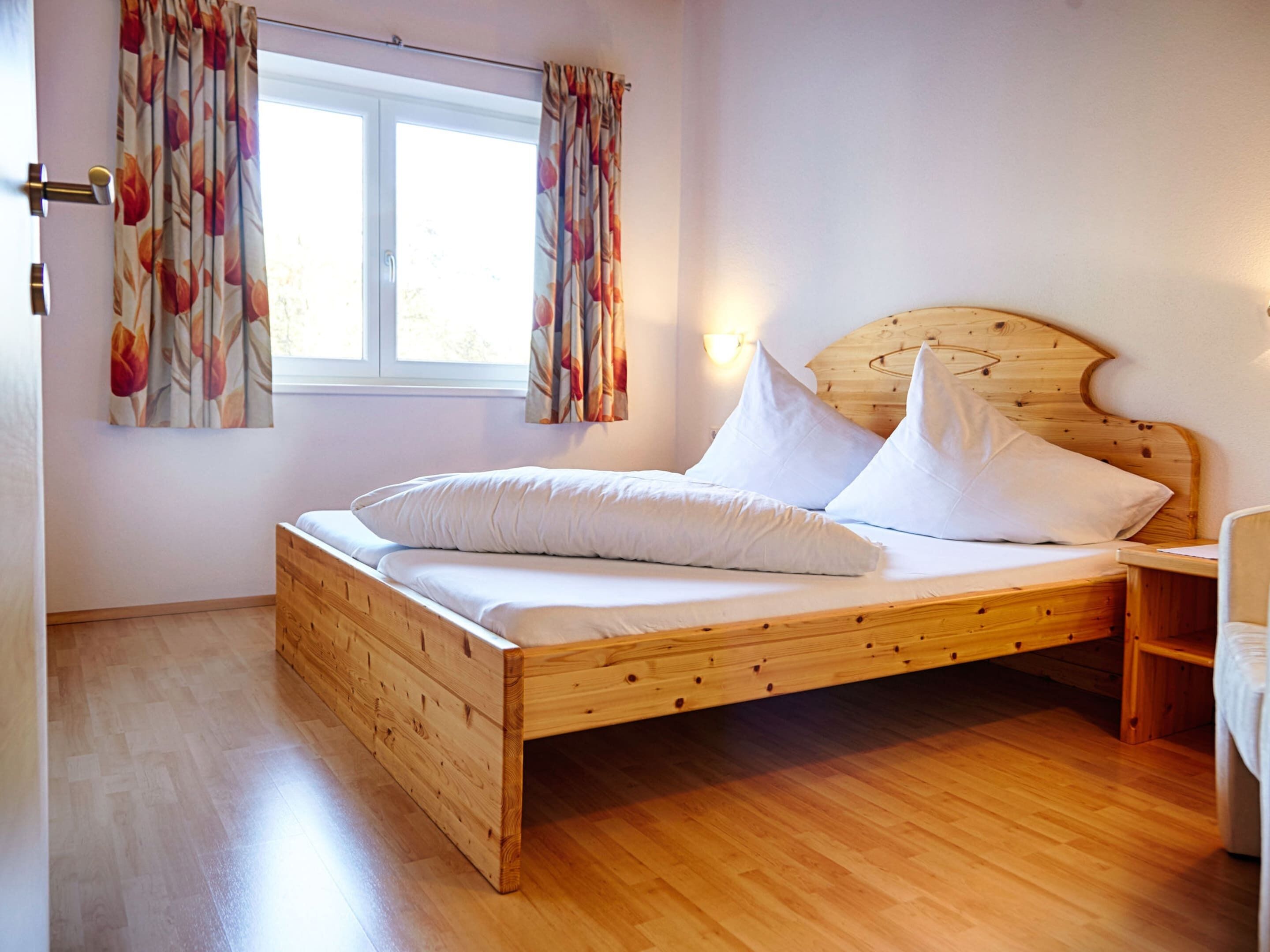 Una cama de madera con ropa de cama blanca en una habitación con suelo de madera y una ventana con cortinas florales.