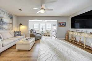 Un salon spacieux avec un grand canapé blanc, une table basse, un ventilateur de plafond et un téléviseur fixé au mur.