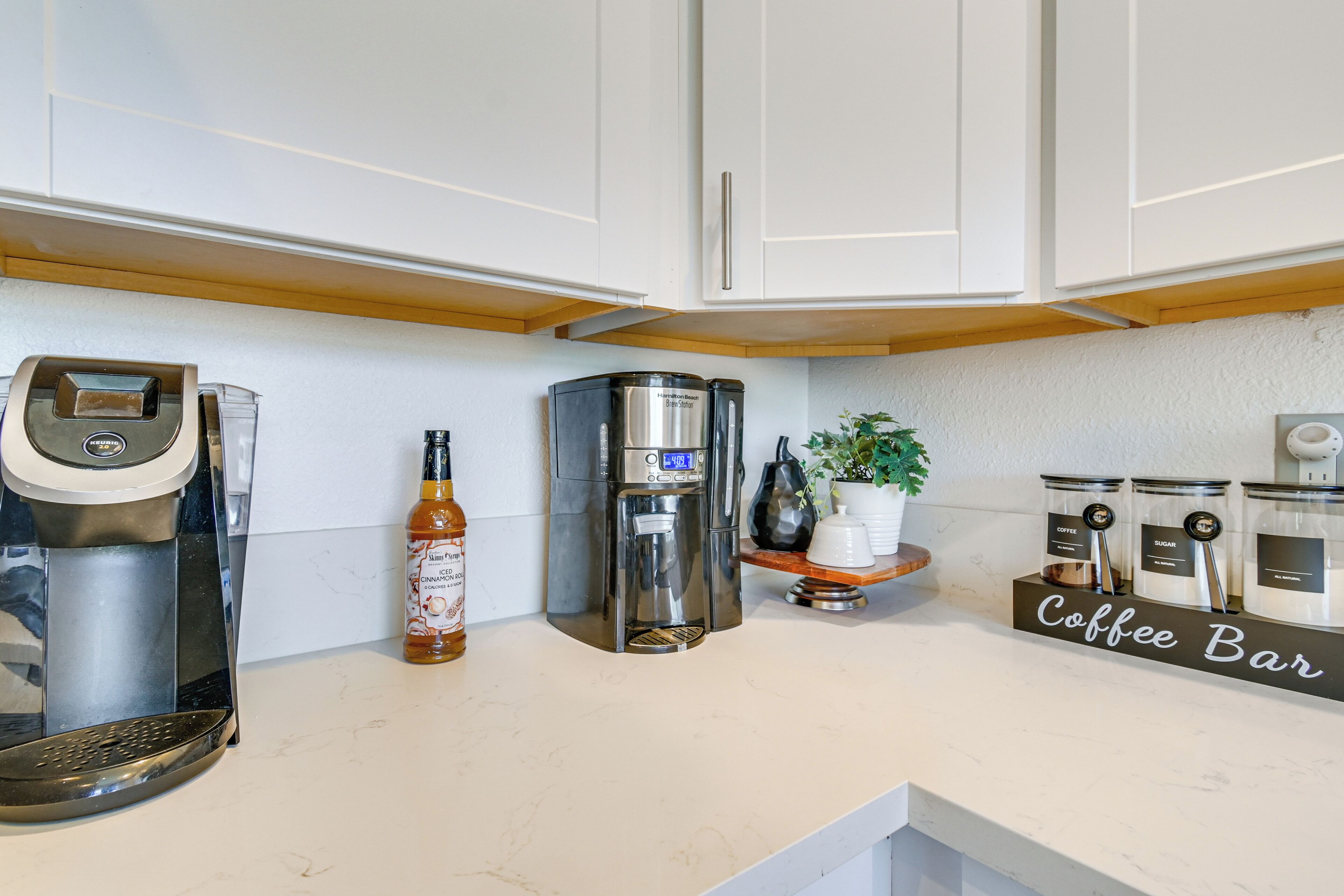 A kitchen with a coffee maker, a bottle of syrup, and a coffee bar sign.