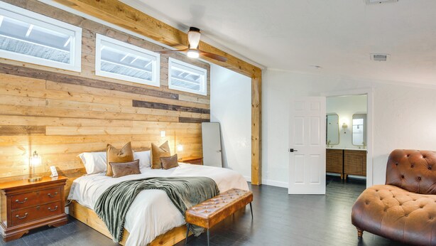 Une chambre à coucher avec une tête de lit en bois, un grand lit avec des oreillers, un banc, une table de chevet et un ventilateur de plafond.
