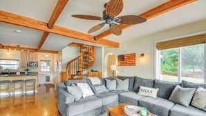 A spacious living area with a gray sectional sofa, wooden ceiling beams, a ceiling fan, a kitchen with a bar area, and a staircase leading to another level. - Bayfront Home w/ Private Beach in Vaughn! (Vaughn)