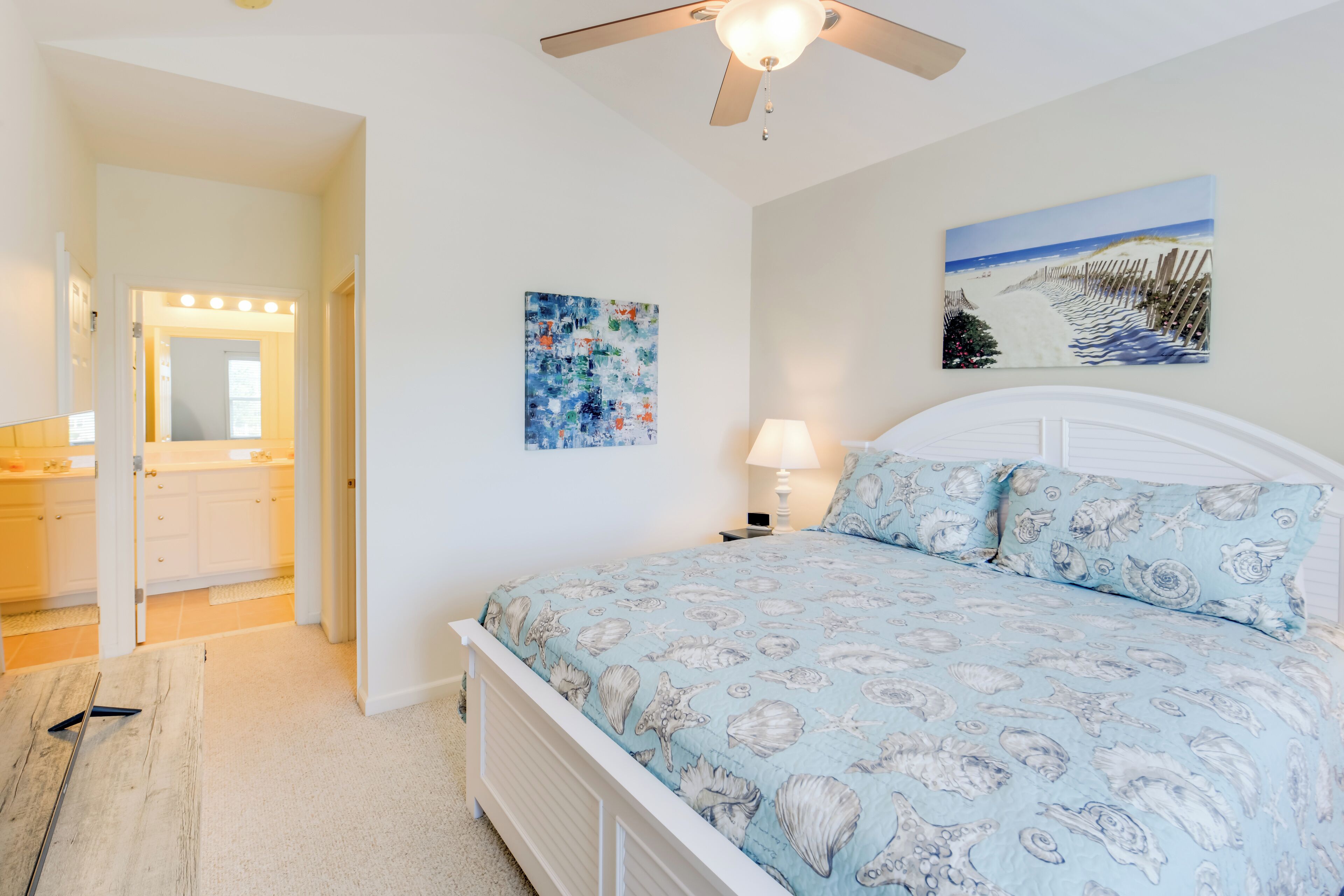 A bedroom with a bed, a ceiling fan, a painting, and a bathroom visible through an open door.