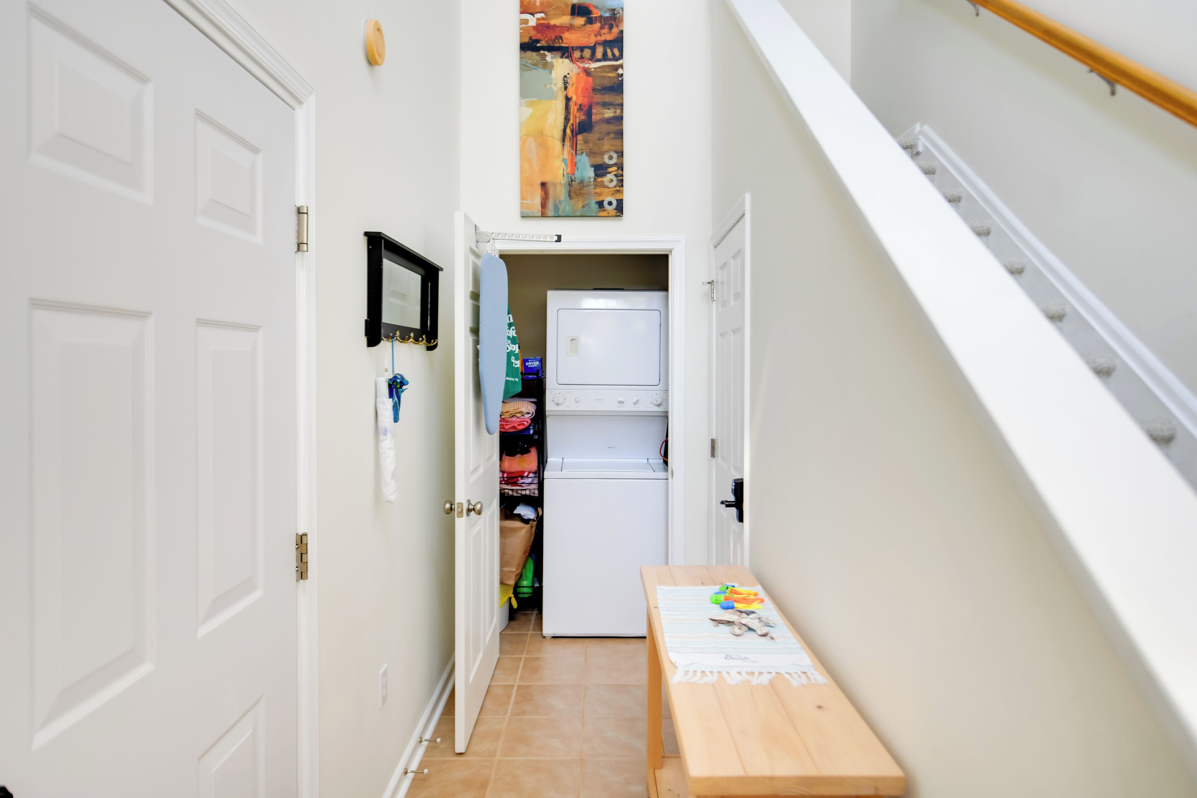 A narrow hallway with a wooden bench, a white door, a refrigerator, and a colorful abstract painting on the wall.
